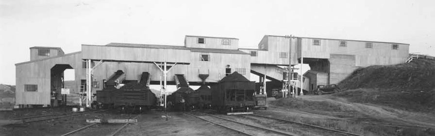 Top: A black and white side shot of the Reliance Tipple, made of corrugated metal. Train cars in various states of being filled with coal sit on tracks beneath portions of the tipple. Bottom: Annie Krek, Christine Cukale, Zabia Mangelos, and Sumiko Hattori in work wear stand at a conveyor built sorting coal from the surrounding rock.
